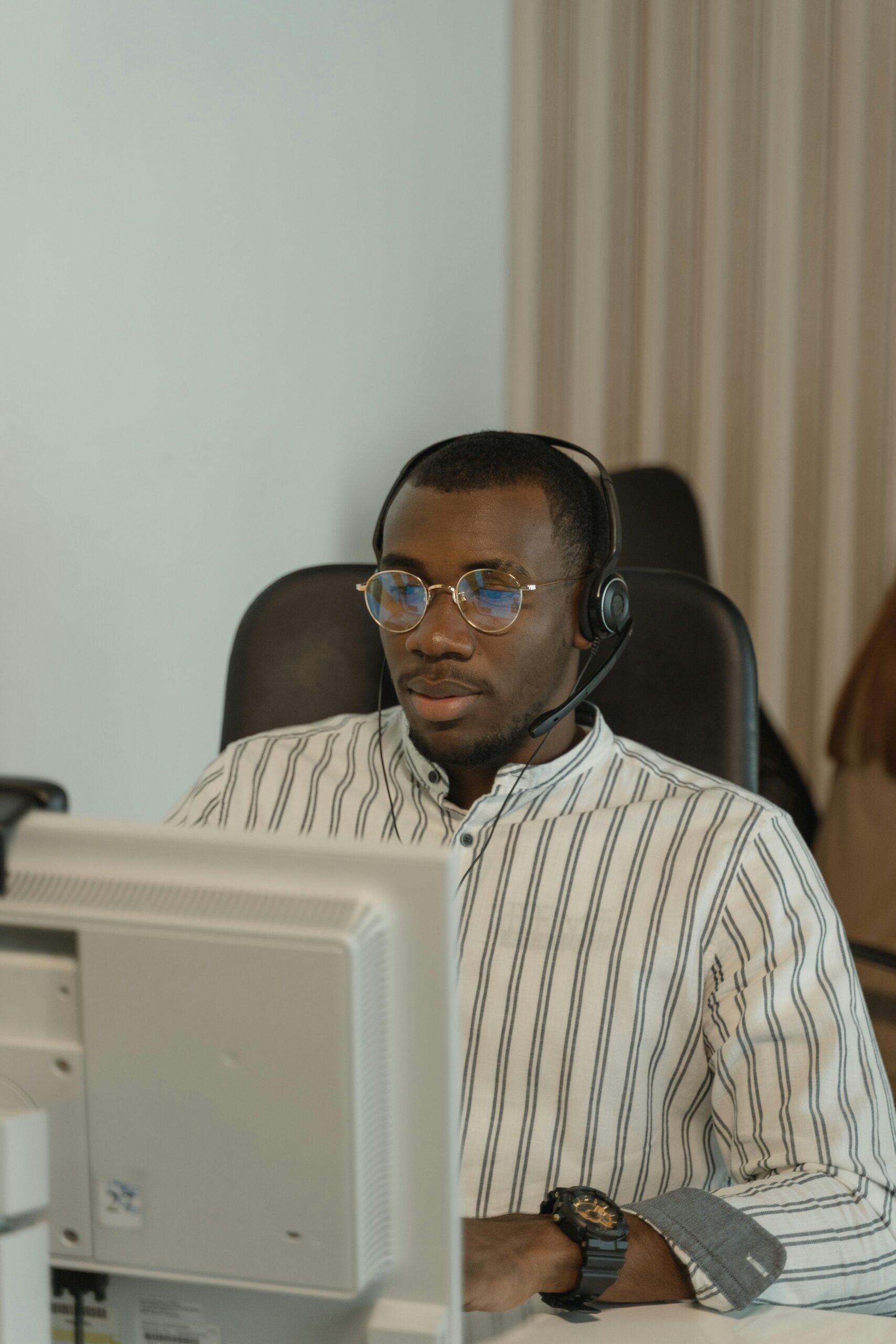 Professional call center agent wearing headset and eyeglasses, working attentively at a computer in an office.